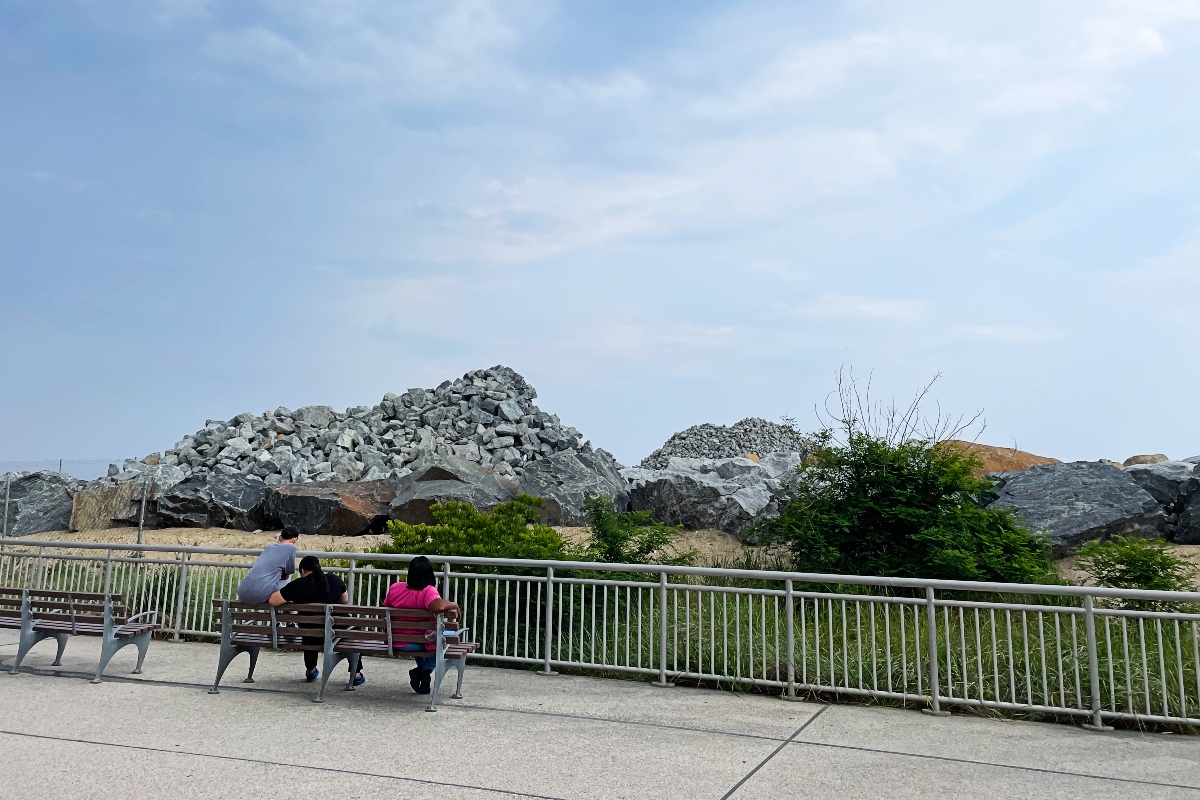 Three people sit on a bench on a beach boardwalk, with a pile of rocks in the distance.