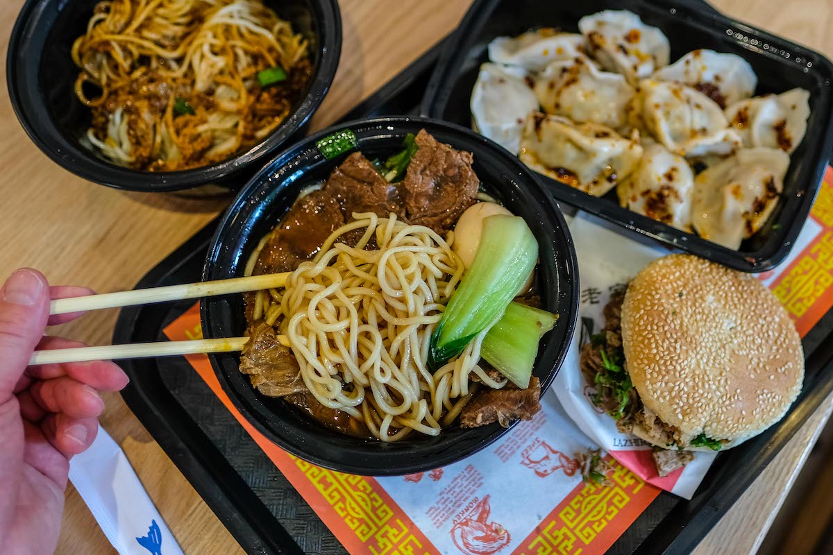 A view of many menu items at Super Taste laid out on a table, with a pair of chopsticks picking up some noodle soup.