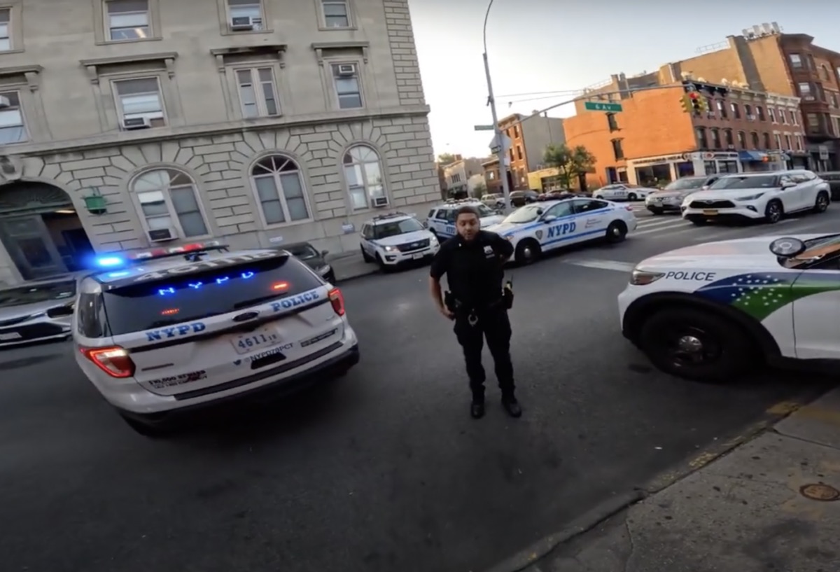 An NYPD SUV attempts to park on the sidewalk outside of a police precinct while an NYPD officer tells a cyclist to get out of their way so they can illegally park.