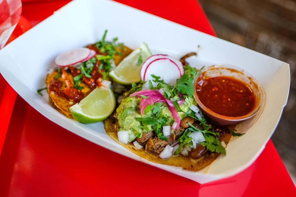 Two birria tacos in a white takeout dish, on a red table.