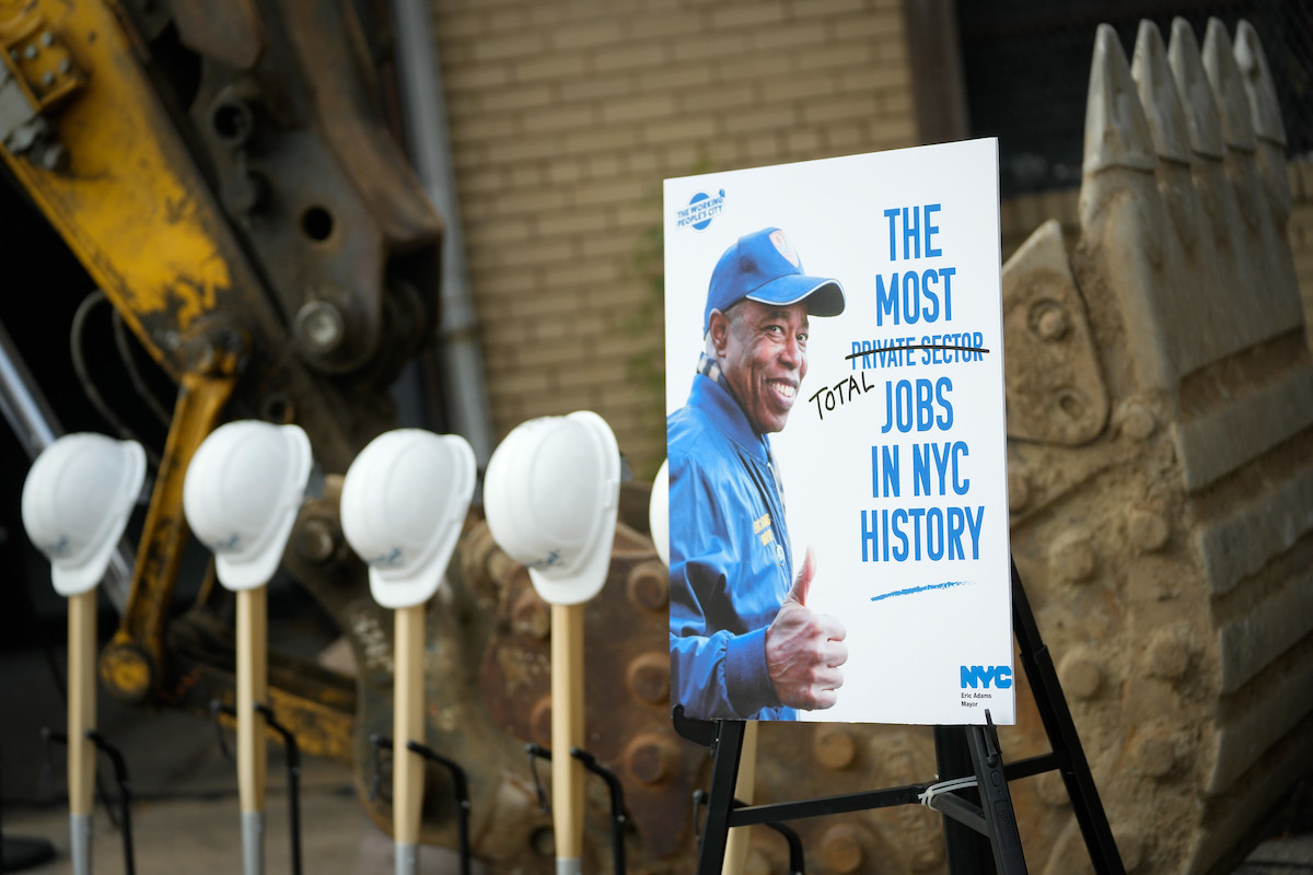 Eric Adams gives a thumbs up on a piece of poster board next to shovels and helmets for a jobs announcment.